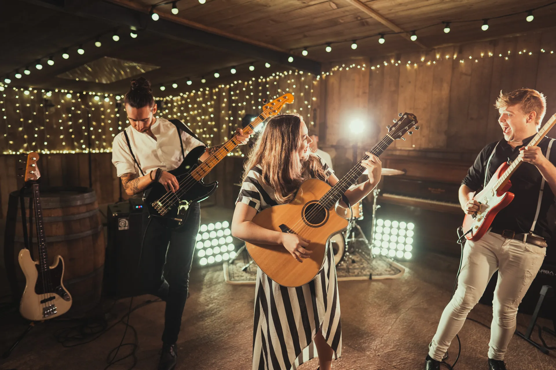 Three musicians playing electric and acoustic guitars in a warmly lit rustic room decorated with string lights.
