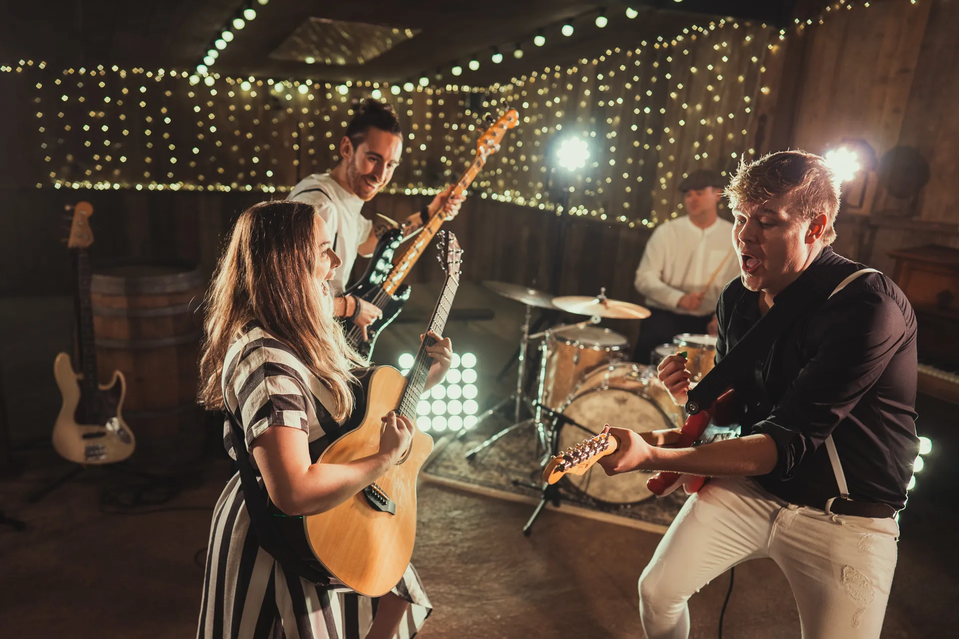 Four-member band performing indoors with string lights, including a woman playing acoustic guitar, two men with electric guitars, and a drummer.