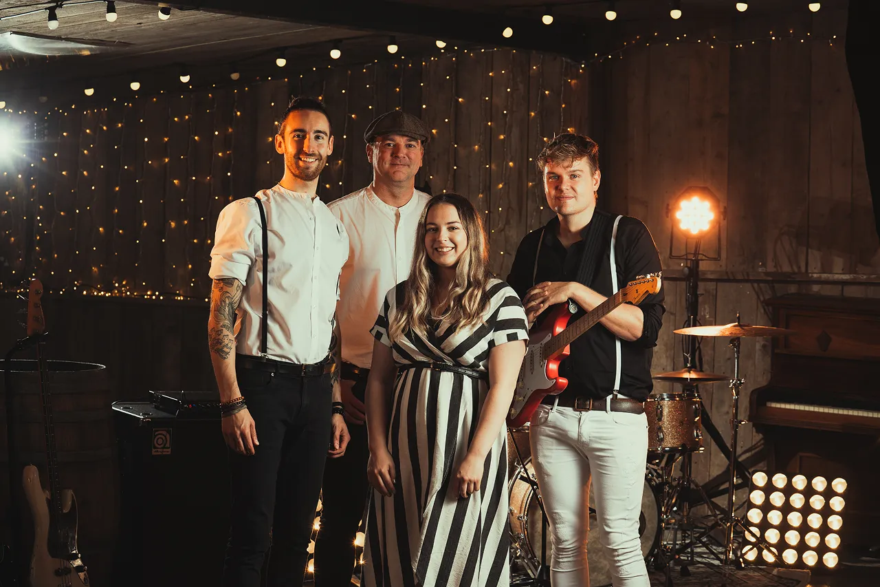 Four musicians posing together on a dimly lit stage with fairy lights and instruments in the background.