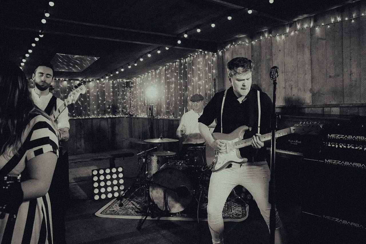Black and white photo of a lively band performing indoors with string lights and wood-paneled walls.