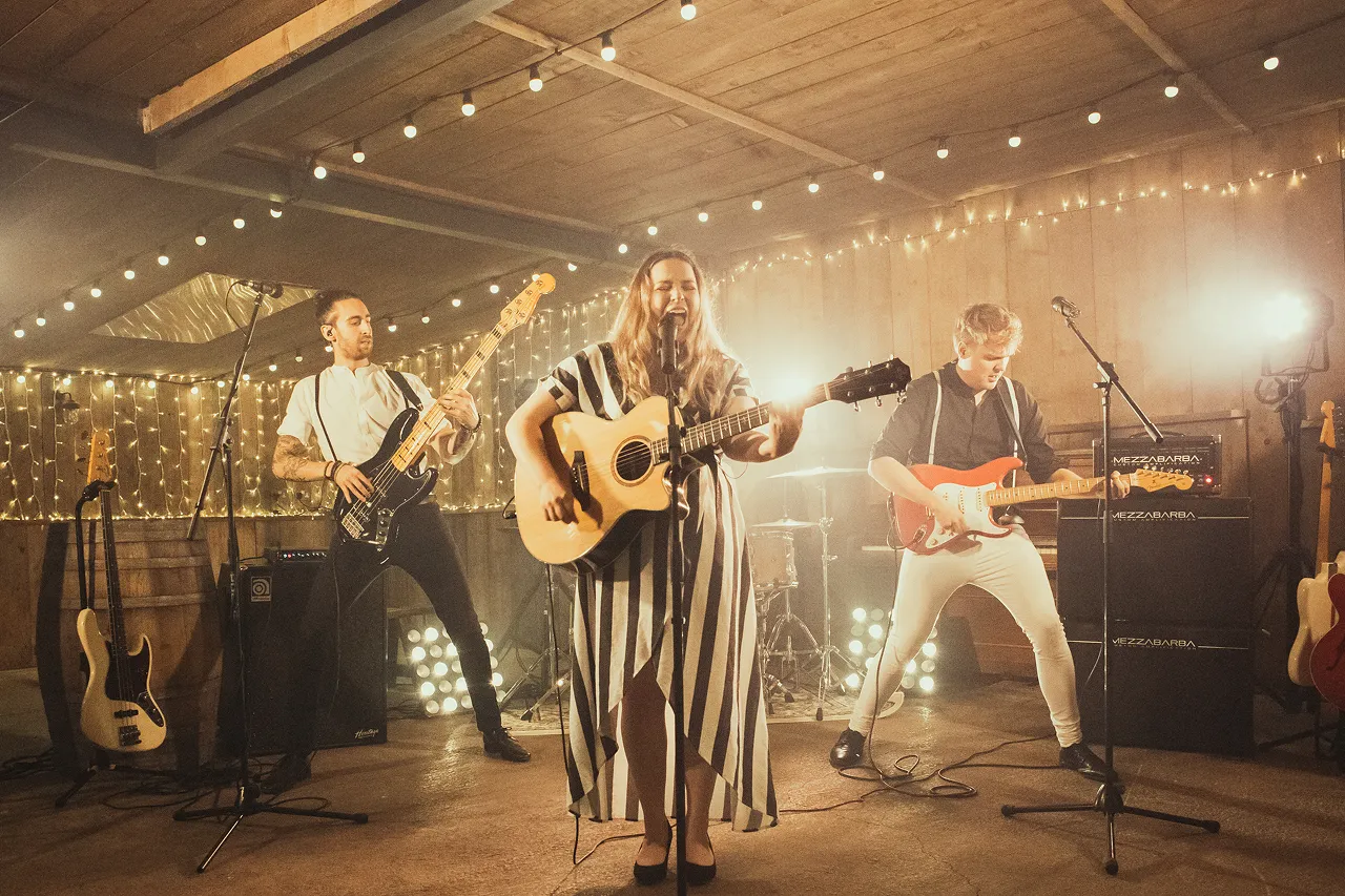 Band performing indoors with string lights overhead and behind, featuring a female singer playing an acoustic guitar and two male guitarists.