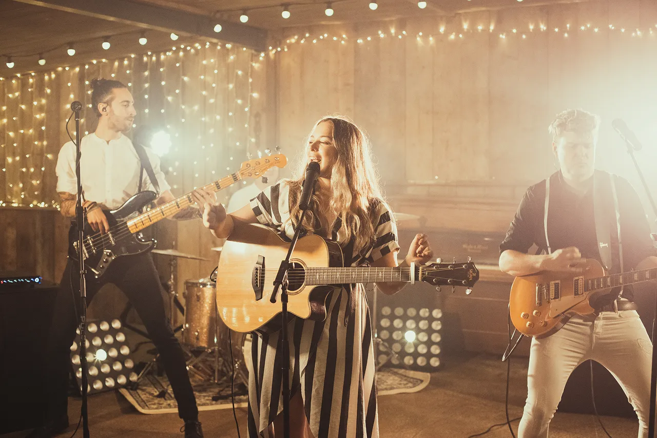 Three musicians performing on stage with guitars and microphone, surrounded by warm string lights.